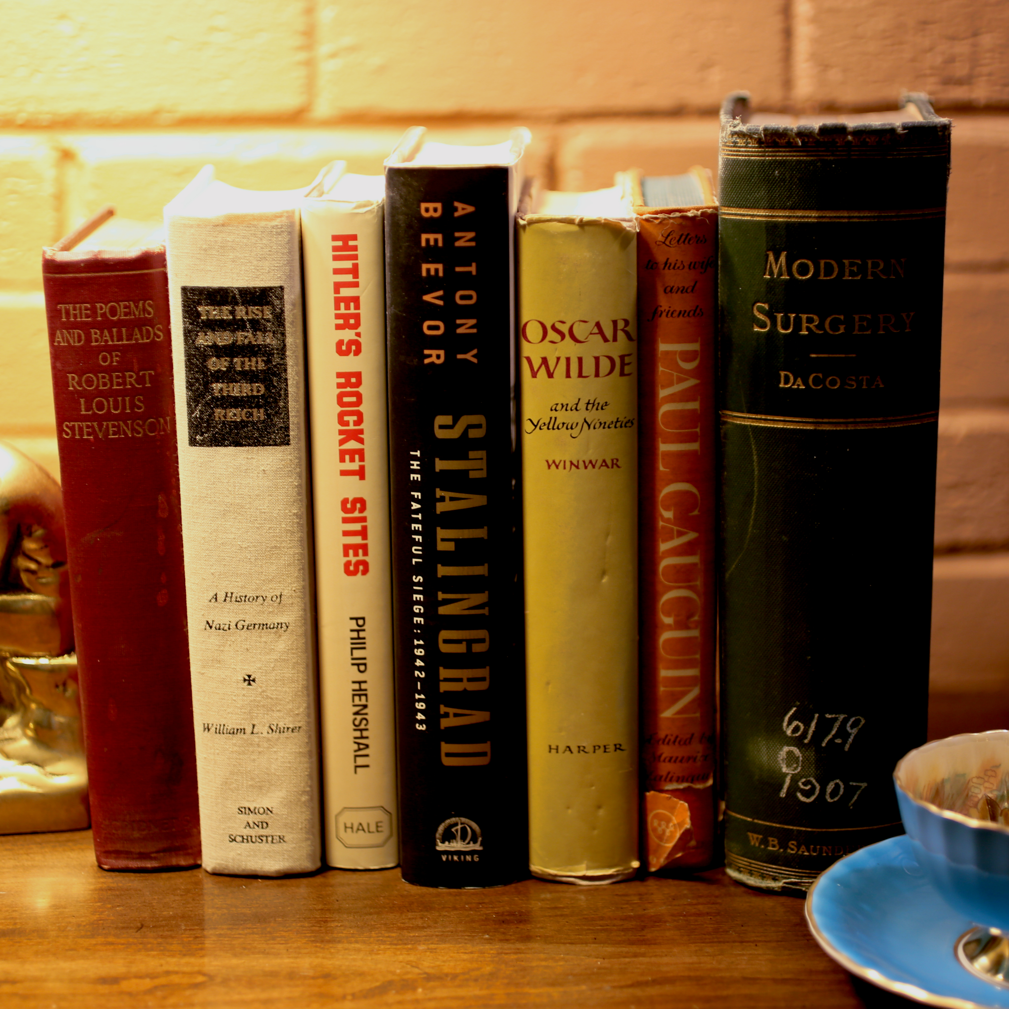 A curated row of vintage and antique books resting on a wooden desk under warm lamplight, featuring well-worn volumes of military history, classic literature, and early 20th-century medical texts, accompanied by a delicate blue teacup.